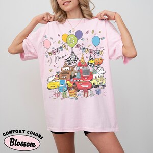 K&ouml;nnte beinhalten: Hellrosa T-Shirt mit einem farbenfrohen Aufdruck von Zeichentrickautos, Luftballons und Geburtstagsdekorationen. Das Shirt tr&auml;gt die Aufschrift "Happy Birthday" und die Zahl "5". Der Text "Name" ist ebenfalls auf dem Shirt. Das Shirt hat auch die Aufschrift "Comfort Colors" und "Blossom".