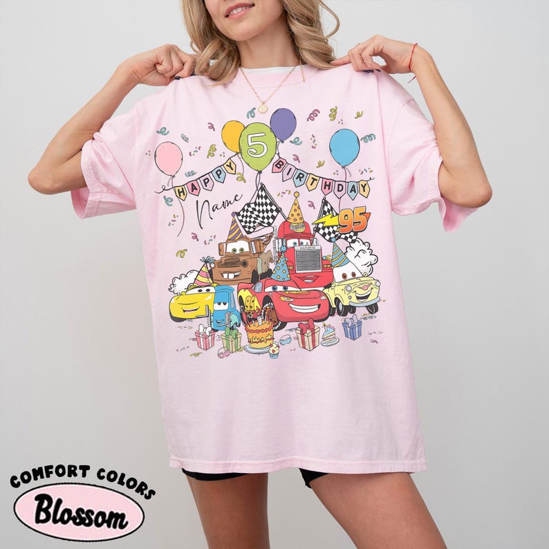 K&ouml;nnte beinhalten: Hellrosa T-Shirt mit einem farbenfrohen Aufdruck von Zeichentrickautos, Luftballons und Geburtstagsdekorationen. Das Shirt tr&auml;gt die Aufschrift "Happy Birthday" und die Zahl "5". Der Text "Name" ist ebenfalls auf dem Shirt. Das Shirt hat auch die Aufschrift "Comfort Colors" und "Blossom".