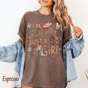 K&ouml;nnte beinhalten: Espressofarbenes T-Shirt mit dem Text "I'm Literally Just a Girl" und Cartoon-Grafiken eines Hufeisens, Cowboyhuts, Stiefeln und eines Pferdes mit einem Cowgirl. Das Shirt hat kurze &Auml;rmel und eine lockere Passform.