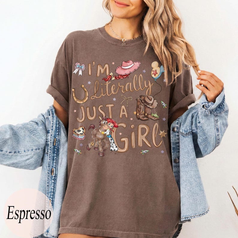 K&ouml;nnte beinhalten: Espressofarbenes T-Shirt mit dem Text "I'm Literally Just a Girl" und Cartoon-Grafiken eines Hufeisens, Cowboyhuts, Stiefeln und eines Pferdes mit einem Cowgirl. Das Shirt hat kurze &Auml;rmel und eine lockere Passform.
