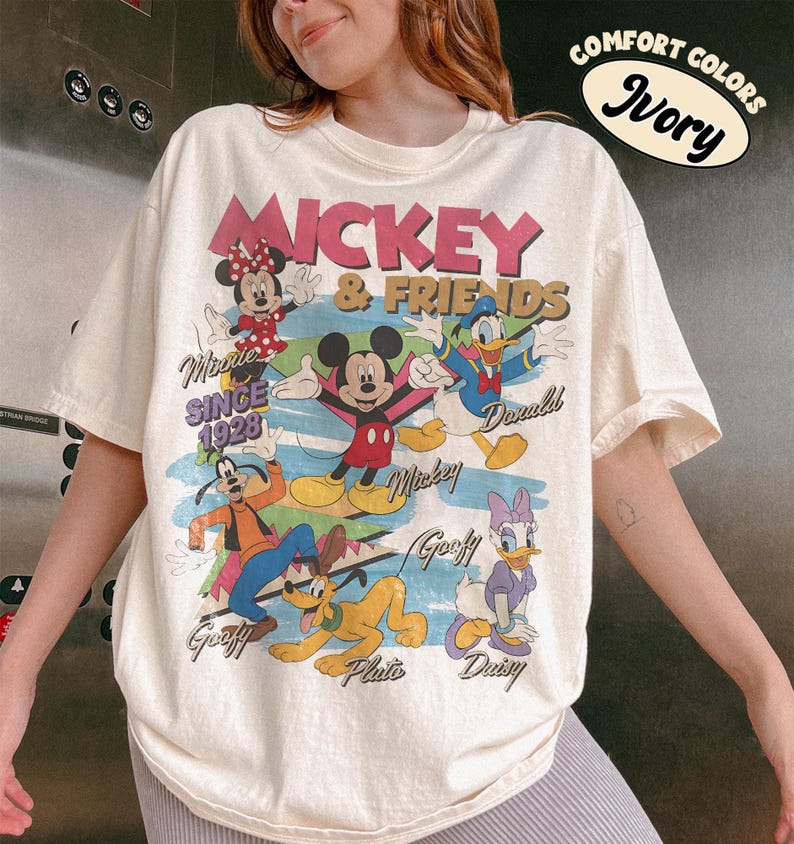 Peut inclure: T-shirt ivoire avec un motif de Mickey Mouse et ses amis. Le design comprend Minnie Mouse, Donald Duck, Dingo, Pluto et Daisy Duck. Le texte "Mickey & Friends Since 1928" est &eacute;galement visible.