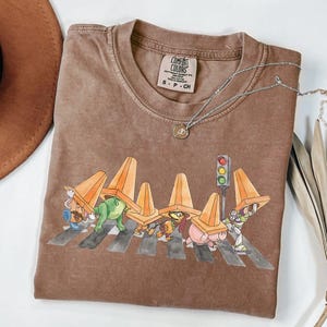 May include: Brown t-shirt with a graphic of cartoon characters crossing a street, each wearing an orange traffic cone. A silver necklace with a pendant is on the shirt. The shirt has the text "Comfort Colors" on the tag.