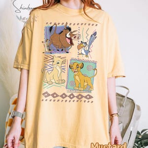 May include: Mustard-colored t-shirt featuring a graphic of characters from The Lion King. The design includes Simba, Timon, Pumbaa, and Zazu in colorful squares. The shirt has a relaxed fit and short sleeves.