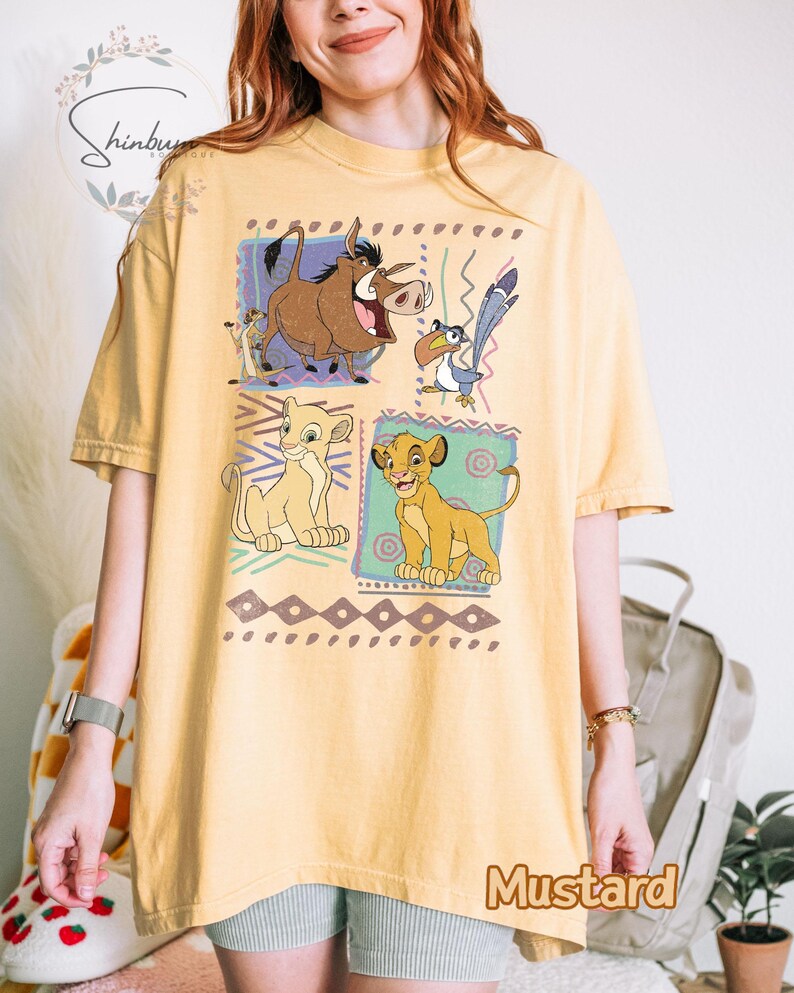 May include: Mustard-colored t-shirt featuring a graphic of characters from The Lion King. The design includes Simba, Timon, Pumbaa, and Zazu in colorful squares. The shirt has a relaxed fit and short sleeves.
