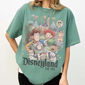May include: A sage green short-sleeve t-shirt featuring a graphic print of Toy Story characters taking a selfie in front of a Disneyland castle. The text Disneyland Est. 1955 is printed in a dark font below the characters on the front of the shirt.