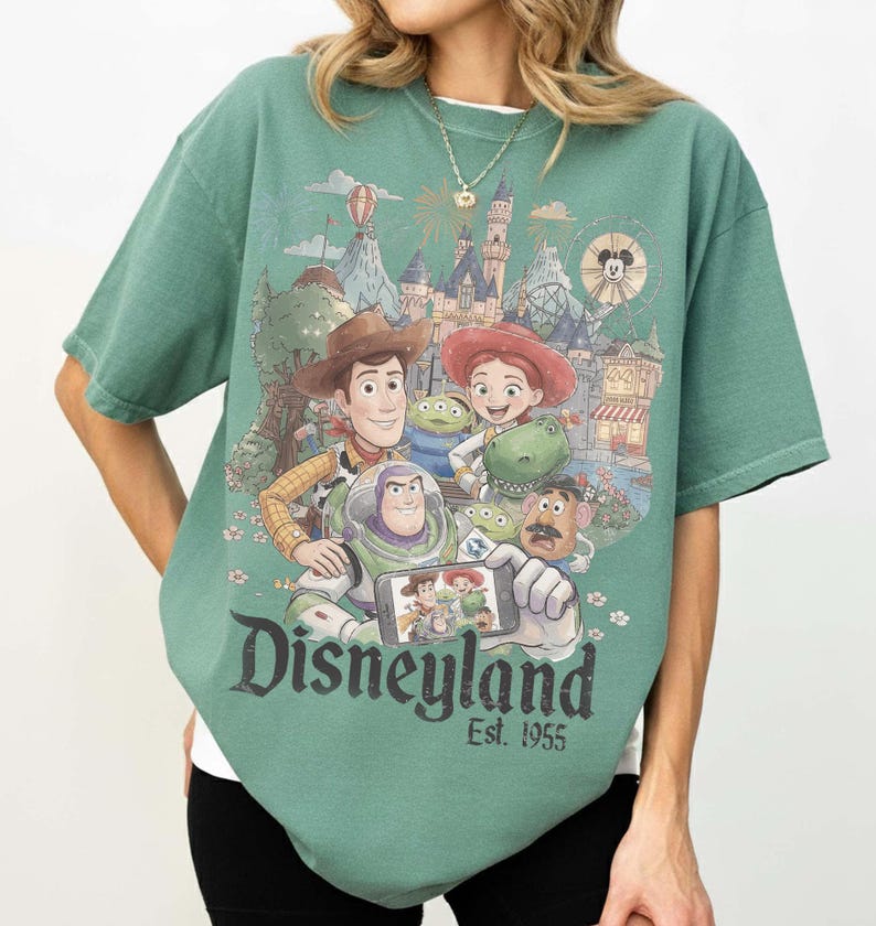 May include: A sage green short-sleeve t-shirt featuring a graphic print of Toy Story characters taking a selfie in front of a Disneyland castle. The text Disneyland Est. 1955 is printed in a dark font below the characters on the front of the shirt.