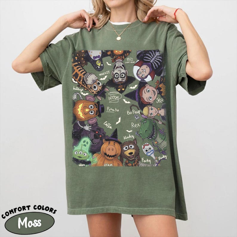 May include: An olive green short-sleeve t-shirt featuring a graphic print of various animated characters in Halloween costumes. The design includes labels for characters like Woody, Buzz Lightyear, Jessie, Bo Peep, Rex, Forky, Slinky, Ham, and Alien.