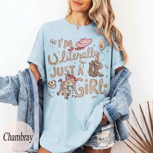 K&ouml;nnte beinhalten: Hellblaues T-Shirt mit dem Text "I'm Literally Just a Girl" und Cowboy-Grafiken. Das Design umfasst ein Hufeisen, Cowboy-Stiefel, ein Pferd und einen rosa Cowboyhut. Das Shirt ist ein l&auml;ssiger Stil mit kurzen &Auml;rmeln.