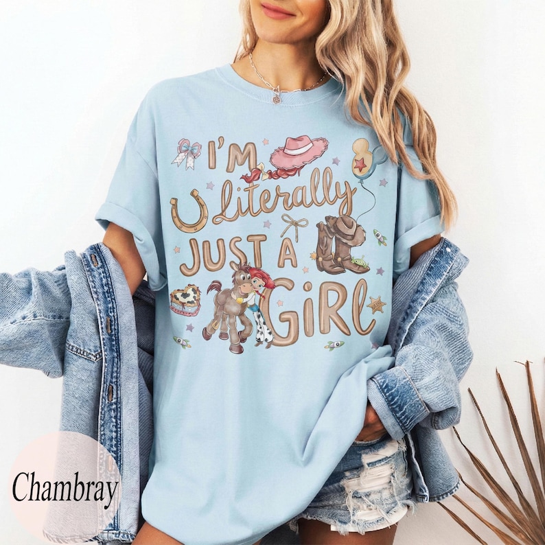 K&ouml;nnte beinhalten: Hellblaues T-Shirt mit dem Text "I'm Literally Just a Girl" und Cowboy-Grafiken. Das Design umfasst ein Hufeisen, Cowboy-Stiefel, ein Pferd und einen rosa Cowboyhut. Das Shirt ist ein l&auml;ssiger Stil mit kurzen &Auml;rmeln.
