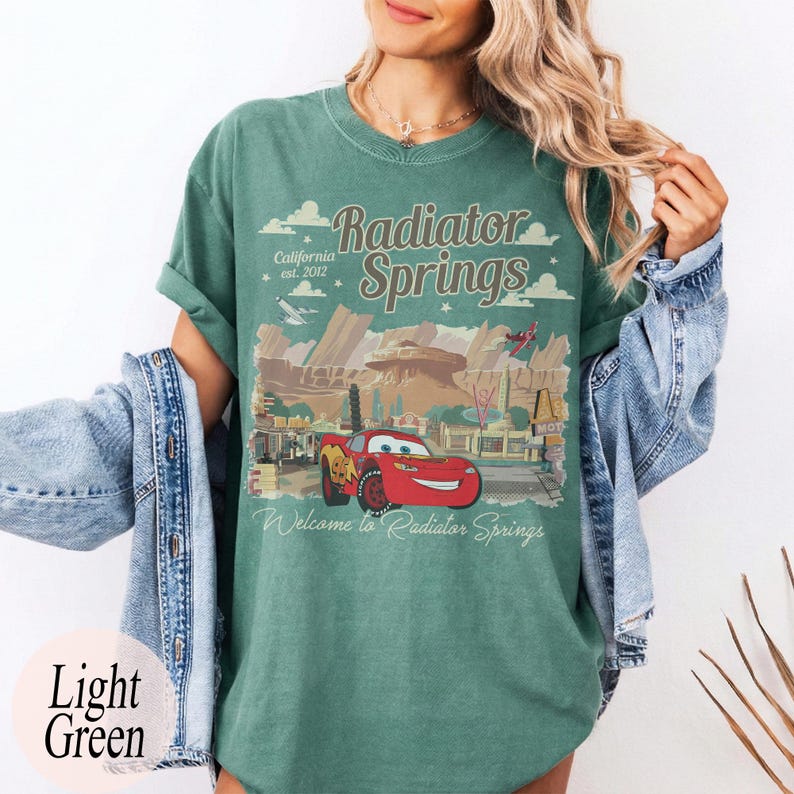 May include: Light green t-shirt with a graphic of Radiator Springs, including a red race car and the text "Radiator Springs" and "Welcome to Radiator Springs". The shirt also has the text "California est. 2012".
