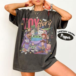 Op de afbeelding: Een antracietgrijs t-shirt met korte mouwen en een grafische print van Toy Story-personages, waaronder Woody, Buzz Lightyear en Rex. Het ontwerp bevat de tekst "TOY story" en "yee haw" met kleurrijke sterren en krabbels. Het shirt is gelabeld als Comfort Colors Pepper.