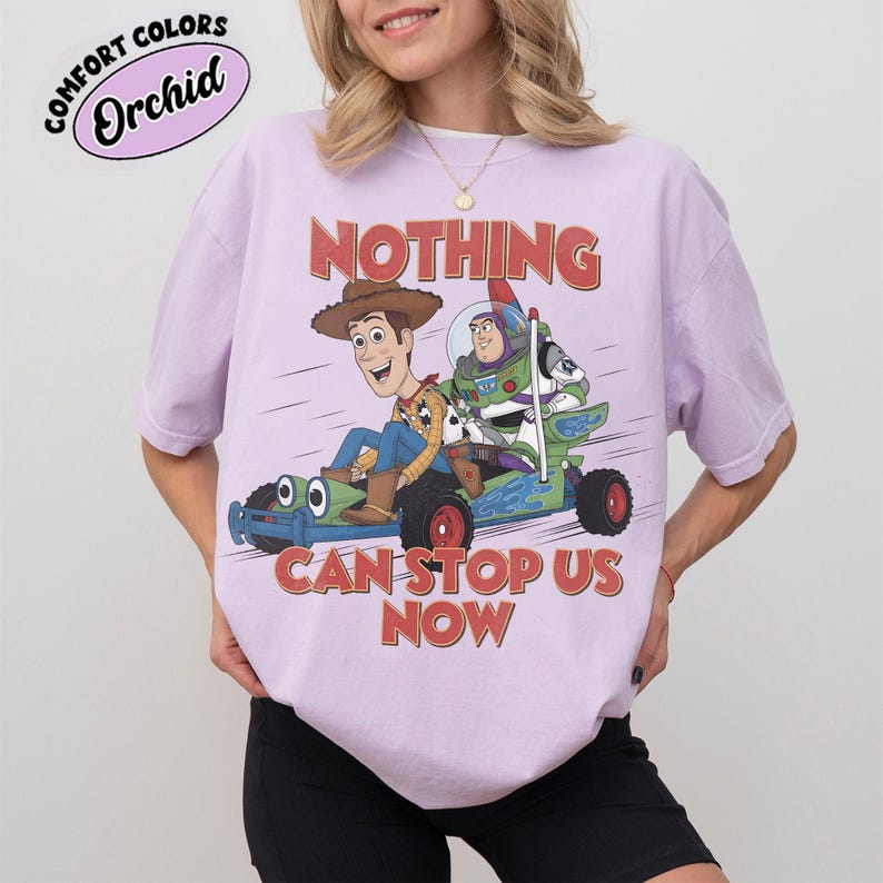 May include: A light purple t-shirt featuring a graphic of Woody and Buzz Lightyear riding a green go-kart. The text above reads NOTHING and below reads CAN STOP US NOW in red letters. A small logo in the top left corner says COMFORT COLORS Orchid.