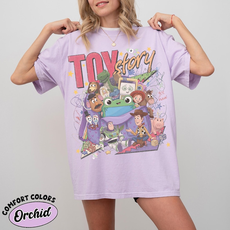 Op de afbeelding: Een orchideekleurig t-shirt met een grote grafische print met de tekst "TOY story" en diverse geanimeerde personages, waaronder een cowboy, space ranger, aardappelfiguur en groene dinosaurus, aangevuld met kleurrijke sterren en krabbels.