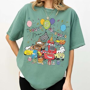 K&ouml;nnte beinhalten: Gr&uuml;nes T-Shirt mit einem farbenfrohen Aufdruck von animierten Autos, Luftballons und dem Text "Happy Birthday" mit einem "5" Ballon. Das Design beinhaltet eine Geburtstagstorte und Geschenkboxen. Das Shirt hat eine lockere Passform.