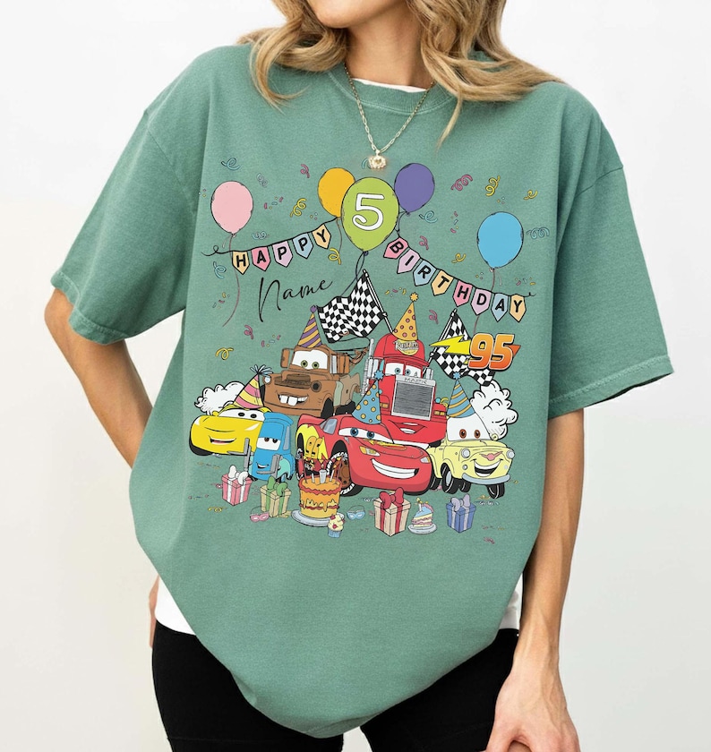 K&ouml;nnte beinhalten: Gr&uuml;nes T-Shirt mit einem farbenfrohen Aufdruck von animierten Autos, Luftballons und dem Text "Happy Birthday" mit einem "5" Ballon. Das Design beinhaltet eine Geburtstagstorte und Geschenkboxen. Das Shirt hat eine lockere Passform.