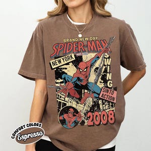 May include: Espresso-coloured t-shirt with a Spider-Man graphic. The design includes the words "BRAND NEW DAY," "SPIDER-MAN," "NEW YORK," and "2008." The graphic shows Spider-Man swinging, with the words "SWING" and "SINTO ACTION."