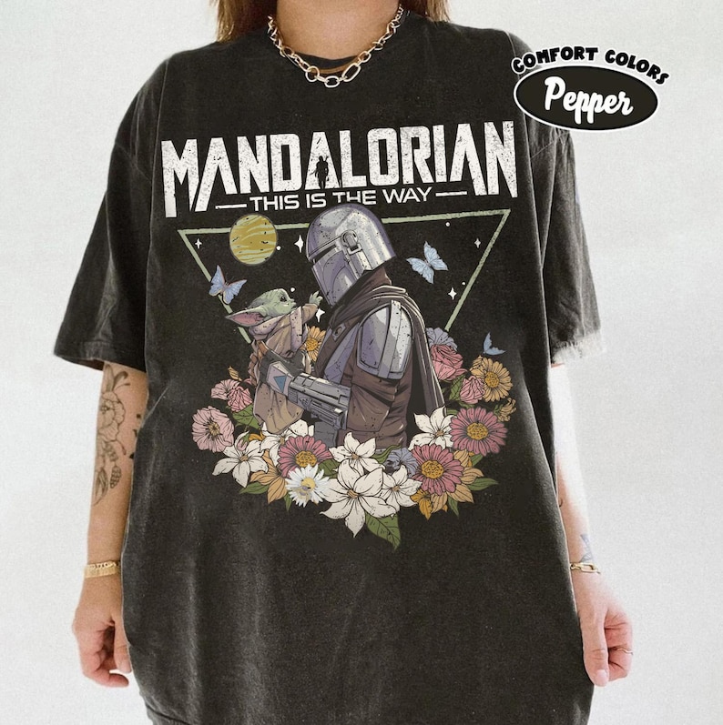 May include: Black t-shirt featuring a graphic of The Mandalorian and Grogu surrounded by flowers and butterflies. The text "MANDALORIAN - THIS IS THE WAY" is printed above the image. The shirt has a distressed look and the words "COMFORT COLORS Pepper" are printed on the upper right.