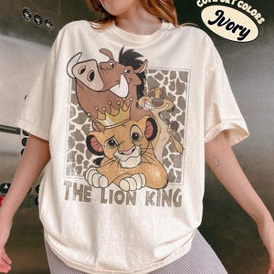 May include: Ivory-colored t-shirt featuring a graphic of characters from The Lion King, including Simba wearing a crown, Pumbaa, and Timon. The text "THE LION KING" is printed below the characters. The shirt also has the text "COMFORT COLORS Ivory".