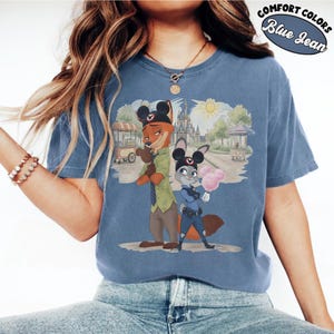 Puede incluir: Camiseta azul vaquero con una ilustraci&oacute;n de dibujos animados de Nick Wilde y Judy Hopps de Zootopia. Los personajes llevan orejas de Mickey Mouse y est&aacute;n de pie frente a un tel&oacute;n de fondo de parque tem&aacute;tico. El texto "Comfort Colors" y "Blue Jean" est&aacute; impreso en la parte superior derecha.