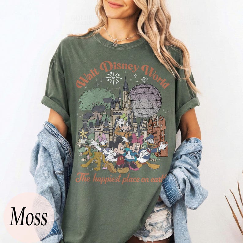 May include: Moss green t-shirt featuring a vintage-style graphic of Walt Disney World with various cartoon characters. The text "Walt Disney World" and "The happiest place on earth" are printed on the front. The shirt has short sleeves and a relaxed fit.
