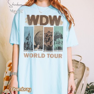 May include: A light blue t-shirt with a graphic print of the Walt Disney World logo and the text "WDW World Tour". The graphic features images of the four theme parks: Magic Kingdom, Epcot Center, Hollywood Studios, and Animal Kingdom.