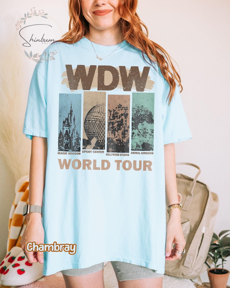 May include: A light blue t-shirt with a graphic print of the Walt Disney World logo and the text "WDW World Tour". The graphic features images of the four theme parks: Magic Kingdom, Epcot Center, Hollywood Studios, and Animal Kingdom.