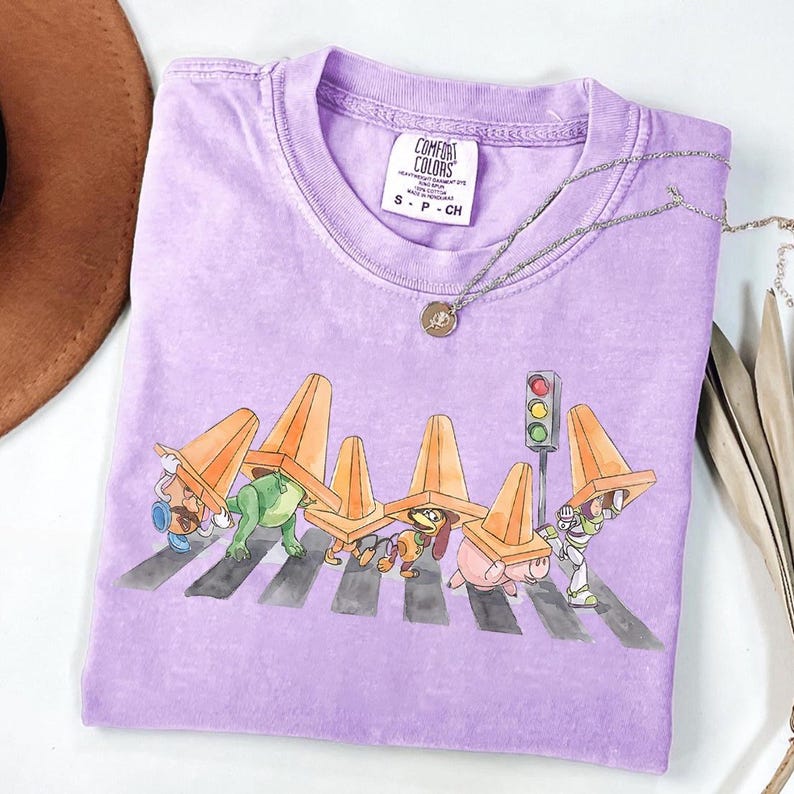 May include: A lilac Comfort Colors t-shirt featuring a graphic of cartoon characters crossing a crosswalk, each wearing an orange traffic cone. A silver necklace with a pendant rests on the shirt.