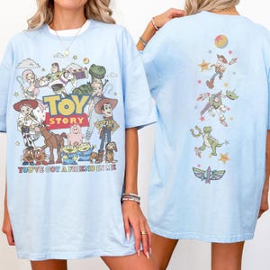 Op de afbeelding: Een lichtblauw t-shirt met een Toy Story-afbeelding op de voor- en achterkant. De voorkant toont personages zoals Woody, Buzz Lightyear en Jessie met de tekst Toy Story en You've Got A Friend In Me. De achterkant toont een verticale rangschikking van personages en iconen.