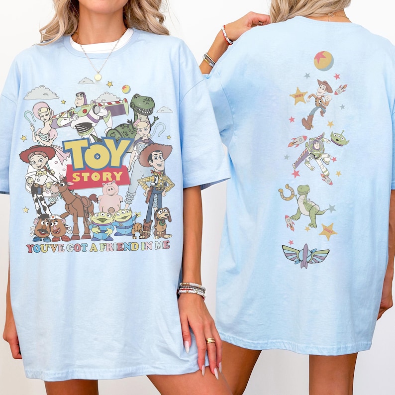 Op de afbeelding: Een lichtblauw t-shirt met een Toy Story-afbeelding op de voor- en achterkant. De voorkant toont personages zoals Woody, Buzz Lightyear en Jessie met de tekst Toy Story en You've Got A Friend In Me. De achterkant toont een verticale rangschikking van personages en iconen.
