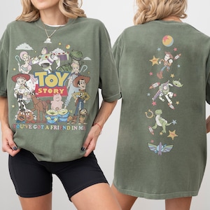 Op de afbeelding: Een olijfgroen t-shirt met Toy Story-personages op de voor- en achterkant. De voorkant toont het Toy Story-logo, personages en de tekst "YOU'VE GOT A FRIEND IN ME". De achterkant toont een verticale rangschikking van personages en sterren.