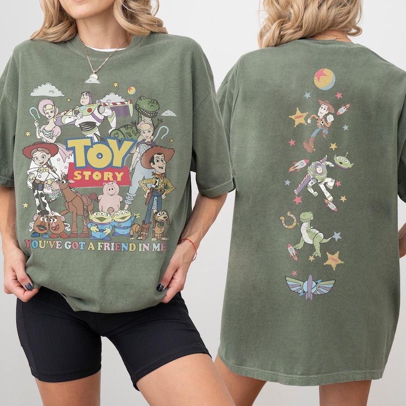 Op de afbeelding: Een olijfgroen t-shirt met Toy Story-personages op de voor- en achterkant. De voorkant toont het Toy Story-logo, personages en de tekst "YOU'VE GOT A FRIEND IN ME". De achterkant toont een verticale rangschikking van personages en sterren.