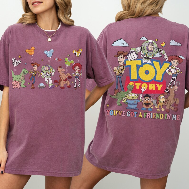 May include: A maroon short-sleeve t-shirt featuring Toy Story characters. The front displays Woody, Buzz Lightyear, and friends with balloons. The back features the Toy Story logo, the characters, and the text "YOU'VE GOT A FRIEND IN ME".
