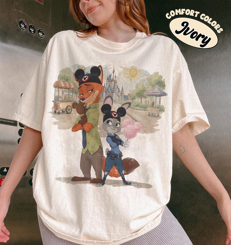 Puede incluir: Camiseta de color marfil con una ilustraci&oacute;n de dibujos animados de Nick Wilde y Judy Hopps de Zootopia, con orejas de Mickey Mouse. El gr&aacute;fico incluye un castillo, un sol y el texto "Comfort Colors Ivory".