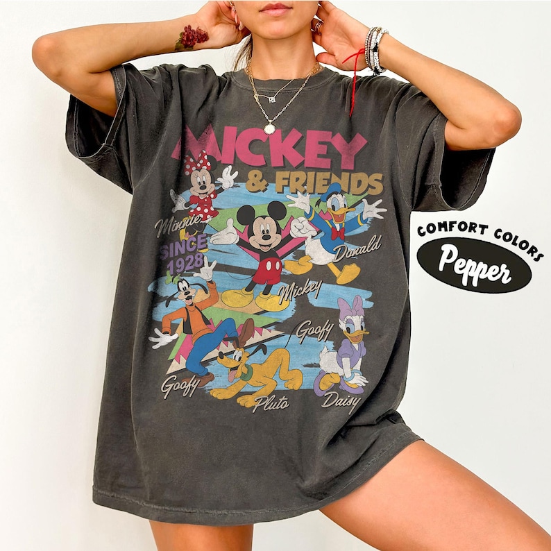 Peut inclure: T-shirt gris fonc&eacute; avec Mickey Mouse et ses amis. Le graphisme comprend Mickey, Minnie, Donald Duck, Dingo, Pluto et Daisy Duck. Le texte comprend "Mickey & Friends" et "Since 1928". Le t-shirt est de Comfort Colors.