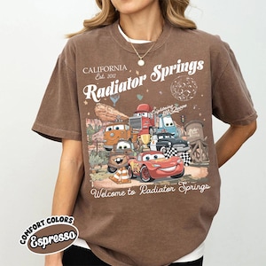以下が含まれることがあります： 映画「カーズ」のキャラクター、ライトニング・マックィーンなどが描かれた茶色のTシャツ。「Radiator Springs, California Est. 2012」の文字と「Welcome to Radiator Springs」の文字もプリントされています。