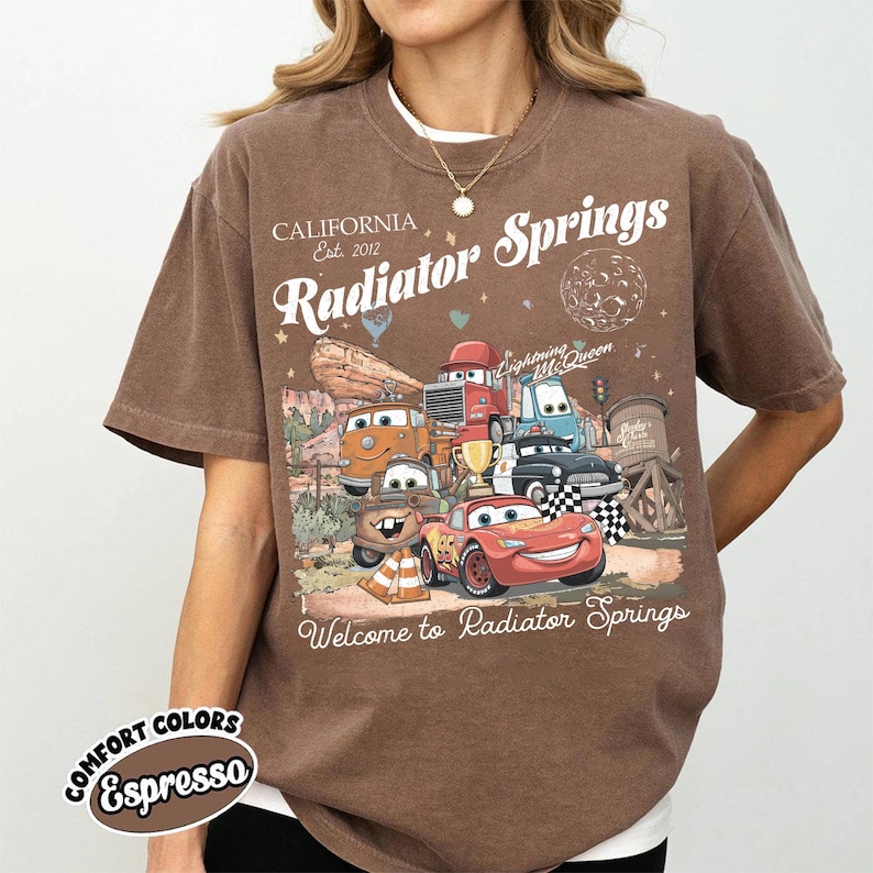 以下が含まれることがあります： 映画「カーズ」のキャラクター、ライトニング・マックィーンなどが描かれた茶色のTシャツ。「Radiator Springs, California Est. 2012」の文字と「Welcome to Radiator Springs」の文字もプリントされています。