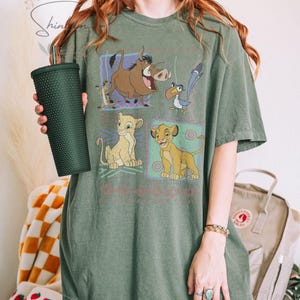 May include: Olive green t-shirt featuring a graphic of characters from The Lion King. The design includes Simba, Timon, Pumbaa, and Zazu in colorful squares. The shirt is a relaxed fit and has short sleeves. A dark green tumbler is held in the left hand.