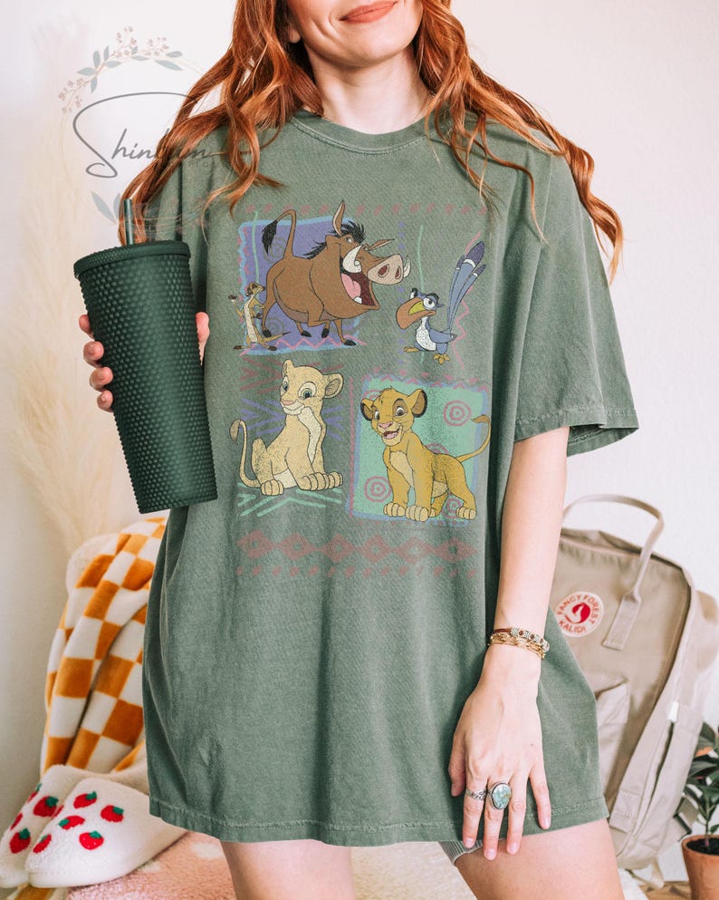 May include: Olive green t-shirt featuring a graphic of characters from The Lion King. The design includes Simba, Timon, Pumbaa, and Zazu in colorful squares. The shirt is a relaxed fit and has short sleeves. A dark green tumbler is held in the left hand.