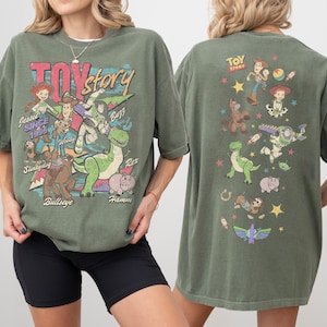 May include: Olive green t-shirt featuring characters from the animated film "Toy Story." The front graphic includes Jessie, Woody, Buzz, Rex, Hamm, Slinky Dog, and Bullseye. The back of the shirt displays additional characters and the film's logo.