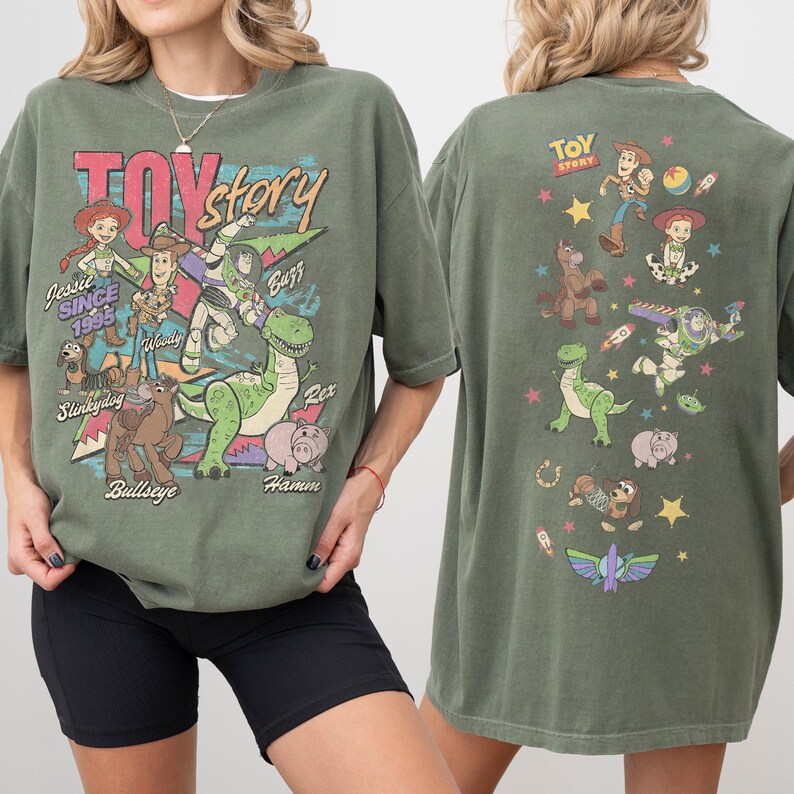 May include: Olive green t-shirt featuring characters from the animated film "Toy Story." The front graphic includes Jessie, Woody, Buzz, Rex, Hamm, Slinky Dog, and Bullseye. The back of the shirt displays additional characters and the film's logo.
