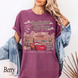May include: Berry-coloured t-shirt featuring a graphic of Lightning McQueen from the movie Cars, with the text "Radiator Springs" and "Welcome to Radiator Springs". The design includes a desert landscape and the text "California est. 2012".