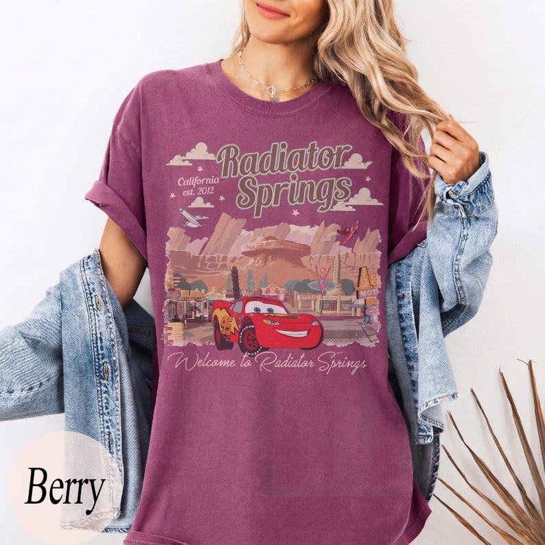 May include: Berry-coloured t-shirt featuring a graphic of Lightning McQueen from the movie Cars, with the text "Radiator Springs" and "Welcome to Radiator Springs". The design includes a desert landscape and the text "California est. 2012".