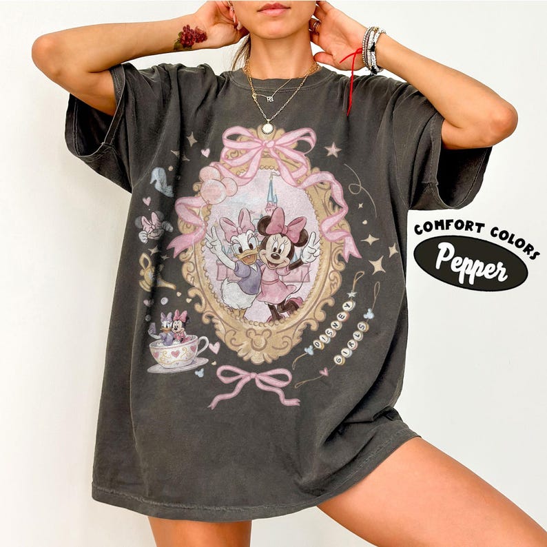 May include: Dark grey t-shirt featuring a framed image of Minnie Mouse and Daisy Duck in pink dresses. The design includes pink bows, stars, and the text "COMFORT COLORS Pepper".
