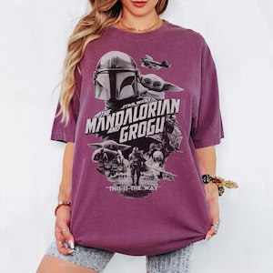 Op de afbeelding: Een bordeauxrood T-shirt met een afbeelding uit de Star Wars-serie, "The Mandalorian and Grogu". Het ontwerp bevat afbeeldingen van de Mandalorian, Grogu en andere personages, met de zin "This is the Way" onderaan.