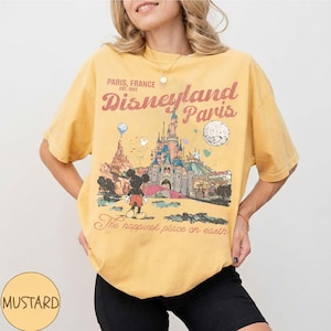 May include: Mustard yellow t-shirt featuring a Disneyland Paris graphic. The design includes the Disneyland Paris castle, Mickey Mouse, and the text "Paris, France" and "The happiest place on earth".