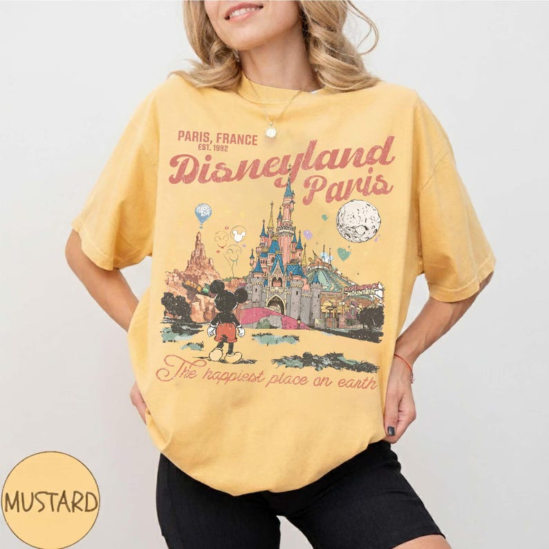 May include: Mustard yellow t-shirt featuring a Disneyland Paris graphic. The design includes the Disneyland Paris castle, Mickey Mouse, and the text "Paris, France" and "The happiest place on earth".