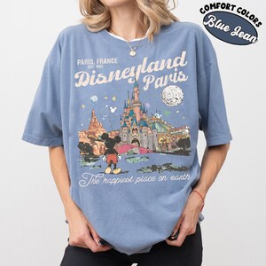 May include: Blue jean-colored t-shirt featuring a graphic of Disneyland Paris. The design includes the castle, Mickey Mouse, and the text "Paris, France" and "The happiest place on earth." The shirt has short sleeves and a crew neck.