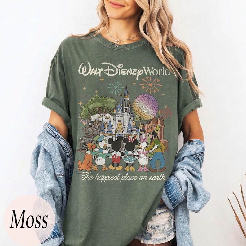Puede incluir: Camiseta verde musgo con un gr&aacute;fico colorido de personajes y lugares emblem&aacute;ticos de Walt Disney World. El texto "Walt Disney World" y "The happiest place on earth" est&aacute;n impresos en la parte delantera. La camiseta tiene mangas cortas y un corte holgado.