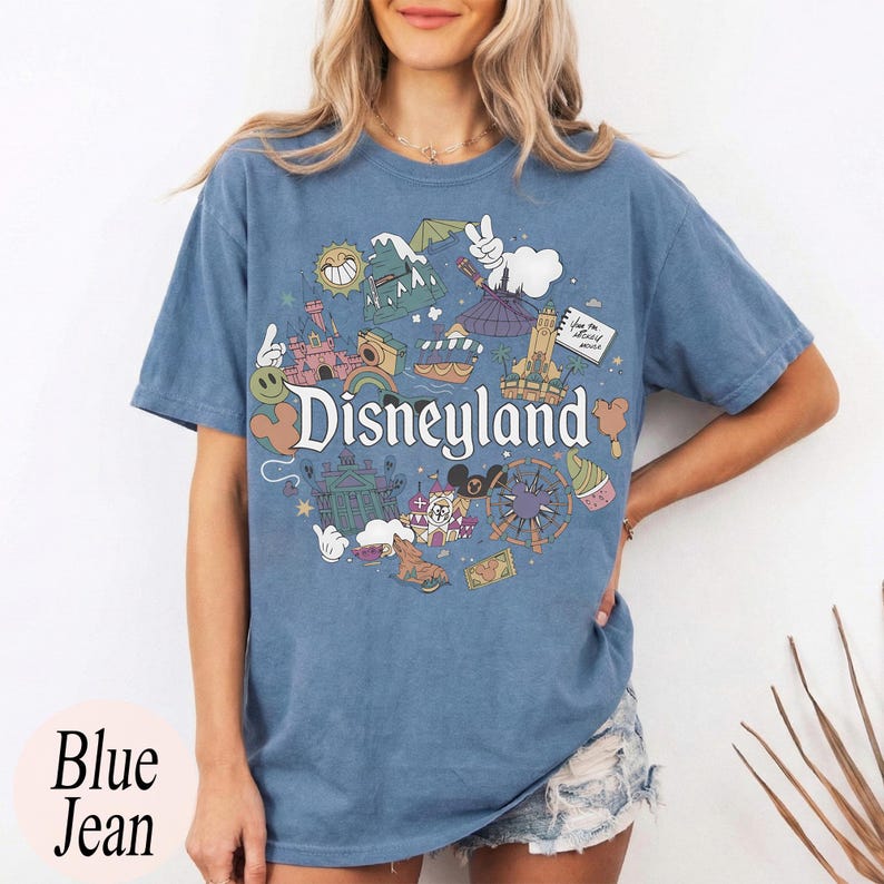 May include: Blue jean-colored t-shirt featuring a colorful Disneyland graphic. The design includes iconic park imagery and the word "Disneyland" in a whimsical font. The shirt has short sleeves and a relaxed fit.