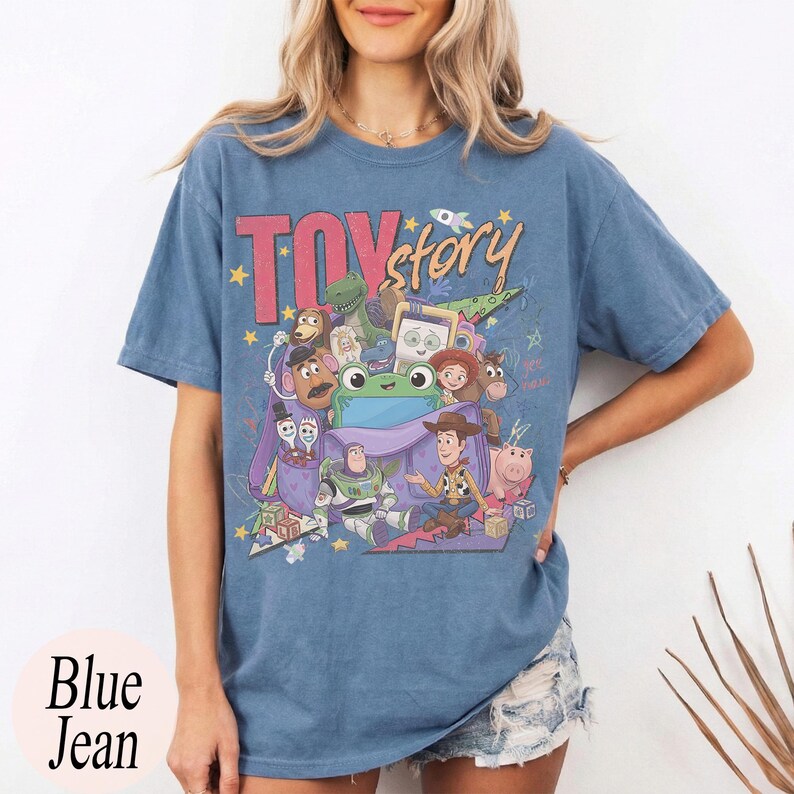 Op de afbeelding: Een blauw spijkerkleurig t-shirt met een grafische print van Toy Story-personages, waaronder Woody en Buzz Lightyear, die uit een paarse tas komen. De tekst Toy Story staat in grote rode en gele letters boven de personages gedrukt.